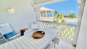 Varanda com mesa branca, cesta de vime e vista para a praia e palmeiras.