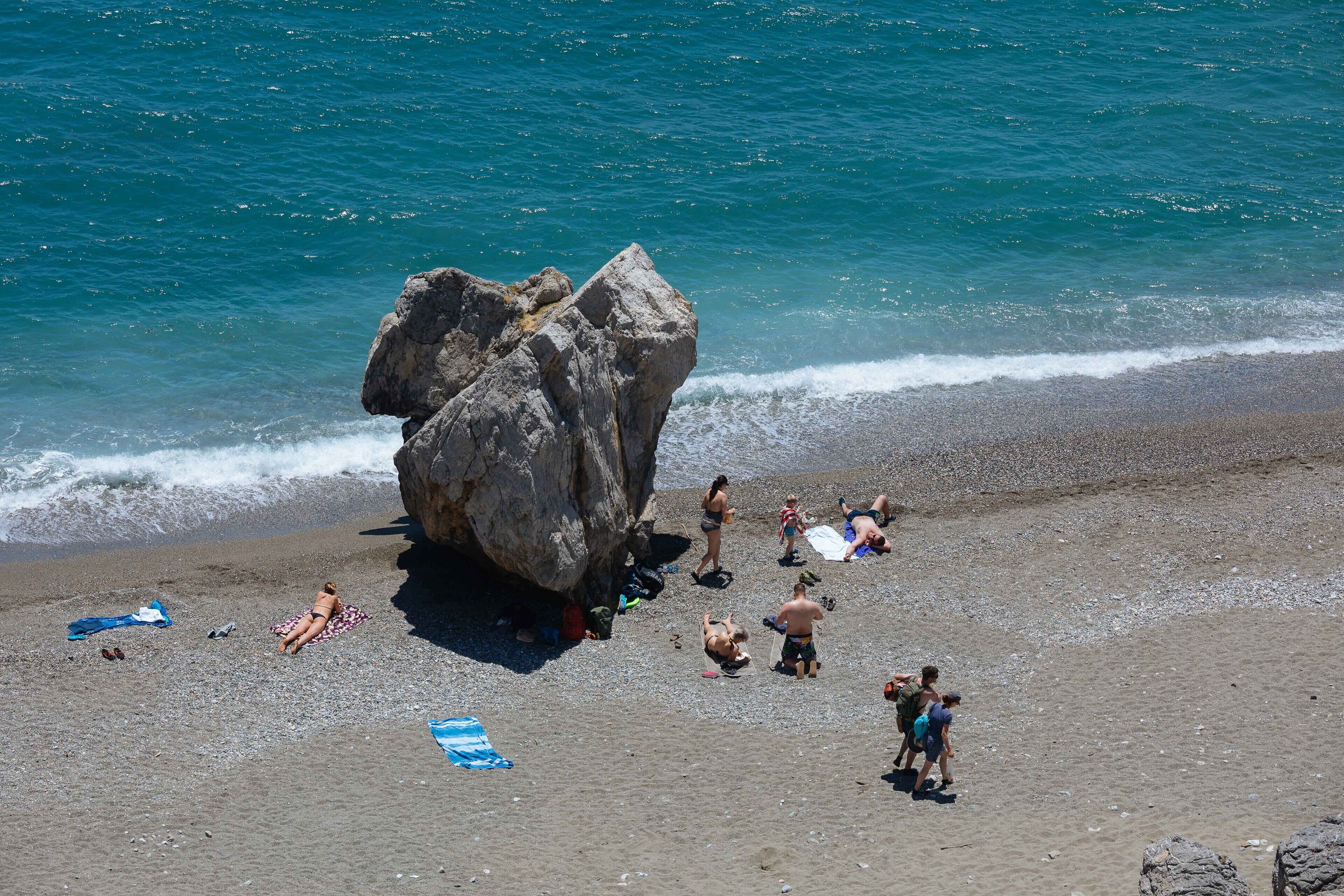 A beach with people sunbathing and walking, a large rock formation, and the ocean in the background.