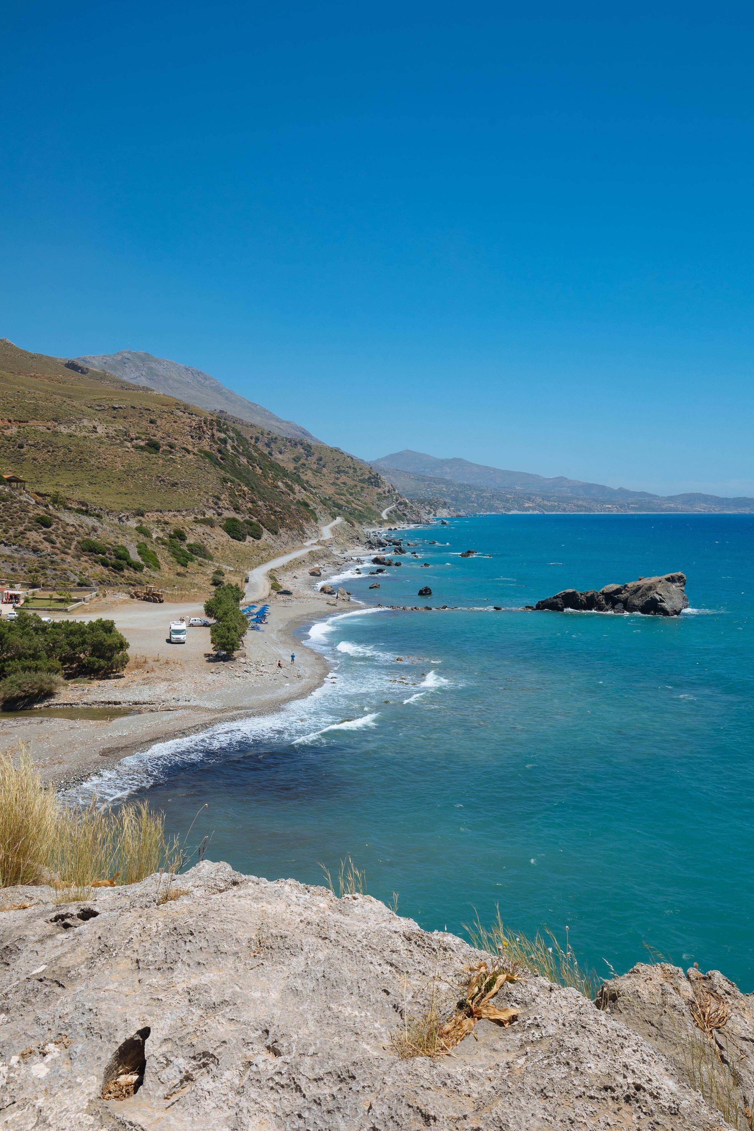 A coastal landscape with a rocky shoreline, clear blue sea, and distant mountains.
