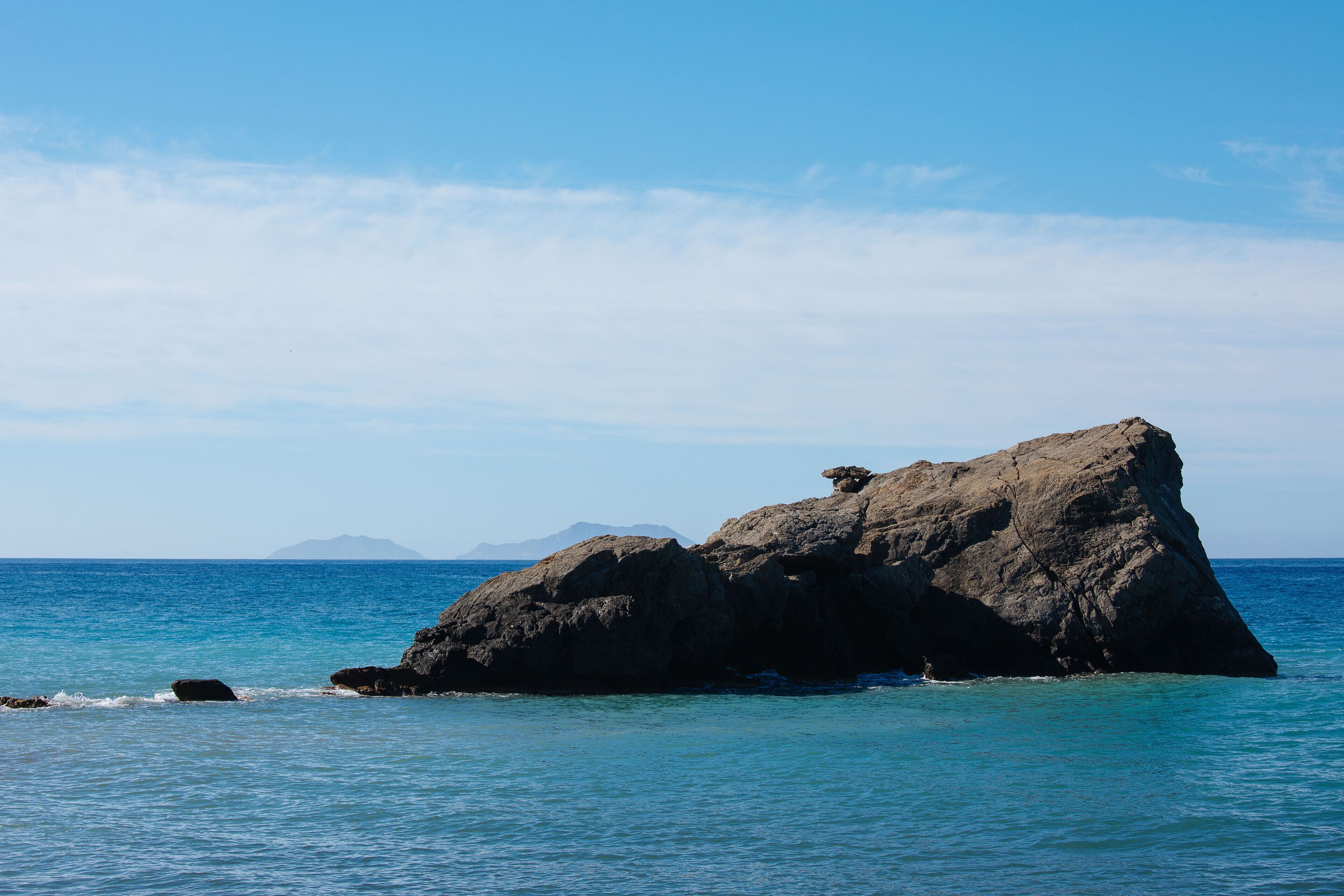 A rocky outcrop in the sea with clear blue water and distant mountains.