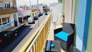 Un balcón con mobiliario de mimbre y una mesa, con vista a una calle con coches y edificios.