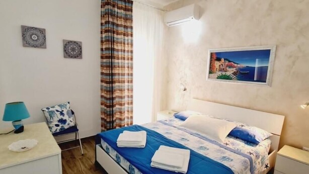 Un dormitorio con cama, mesita de noche, lámpara, silla y un cuadro en la pared.