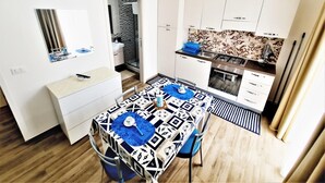A compact kitchen with white cabinets, a stove, and a sink. There's a dining table with a patterned tablecloth and chairs. - Appartamento lu Sule (Porto cesareo)