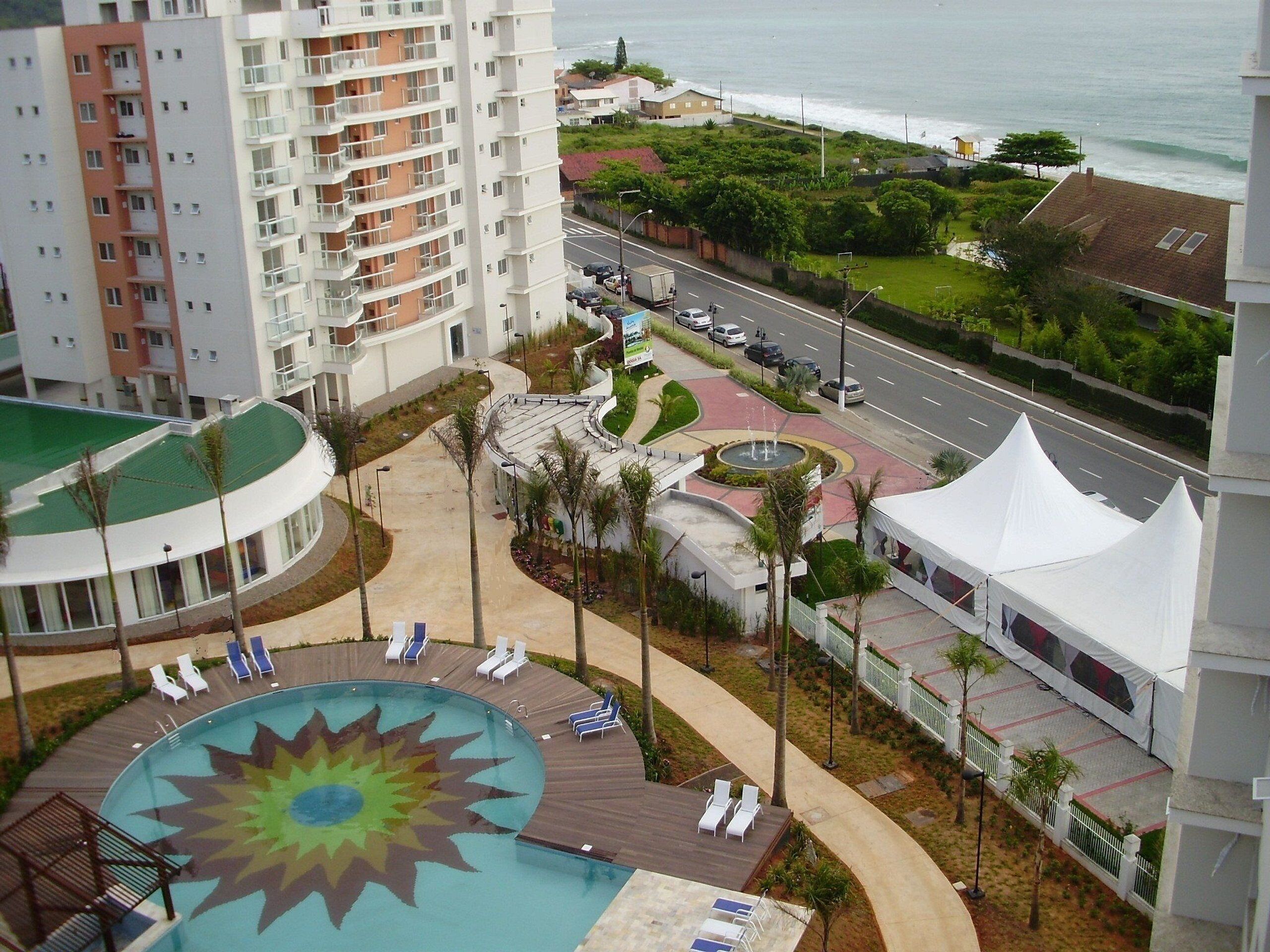 A hotel with a pool featuring a sun design, a road with parked cars, and a view of the ocean.