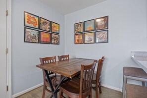 A dining area with a wooden table and chairs, a white wall, and framed pictures. - Boardwalk Shores C 3 Bedroom Duplex by RedAwning (Atlantic Beach)
