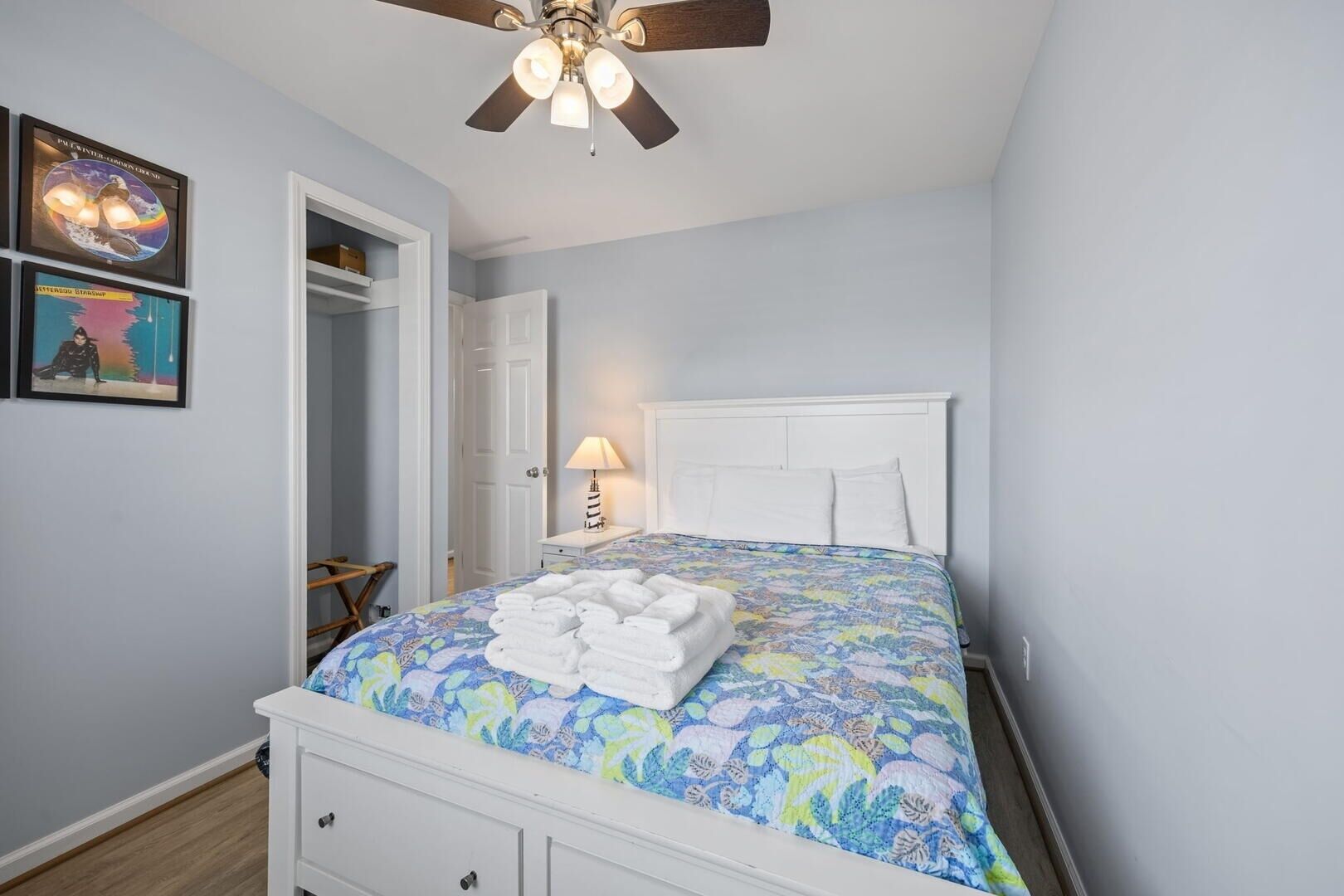 Quarto com cama, ventilador de teto, mesa de cabeceira, abajur e duas imagens emolduradas na parede.