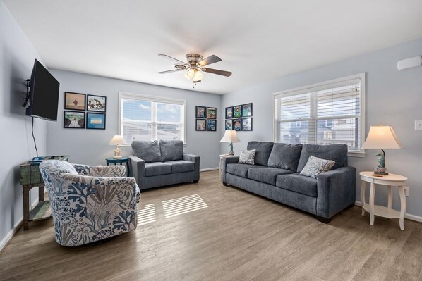 A living room with a grey sofa, a patterned armchair, a ceiling fan, and artwork on the walls. - Boardwalk Shores C 3 Bedroom Duplex by RedAwning (Atlantic Beach)
