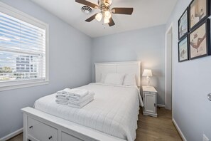 A bedroom with a bed, a ceiling fan, a nightstand with a lamp, and a window with blinds. - Boardwalk Shores C 3 Bedroom Duplex by RedAwning (Atlantic Beach)