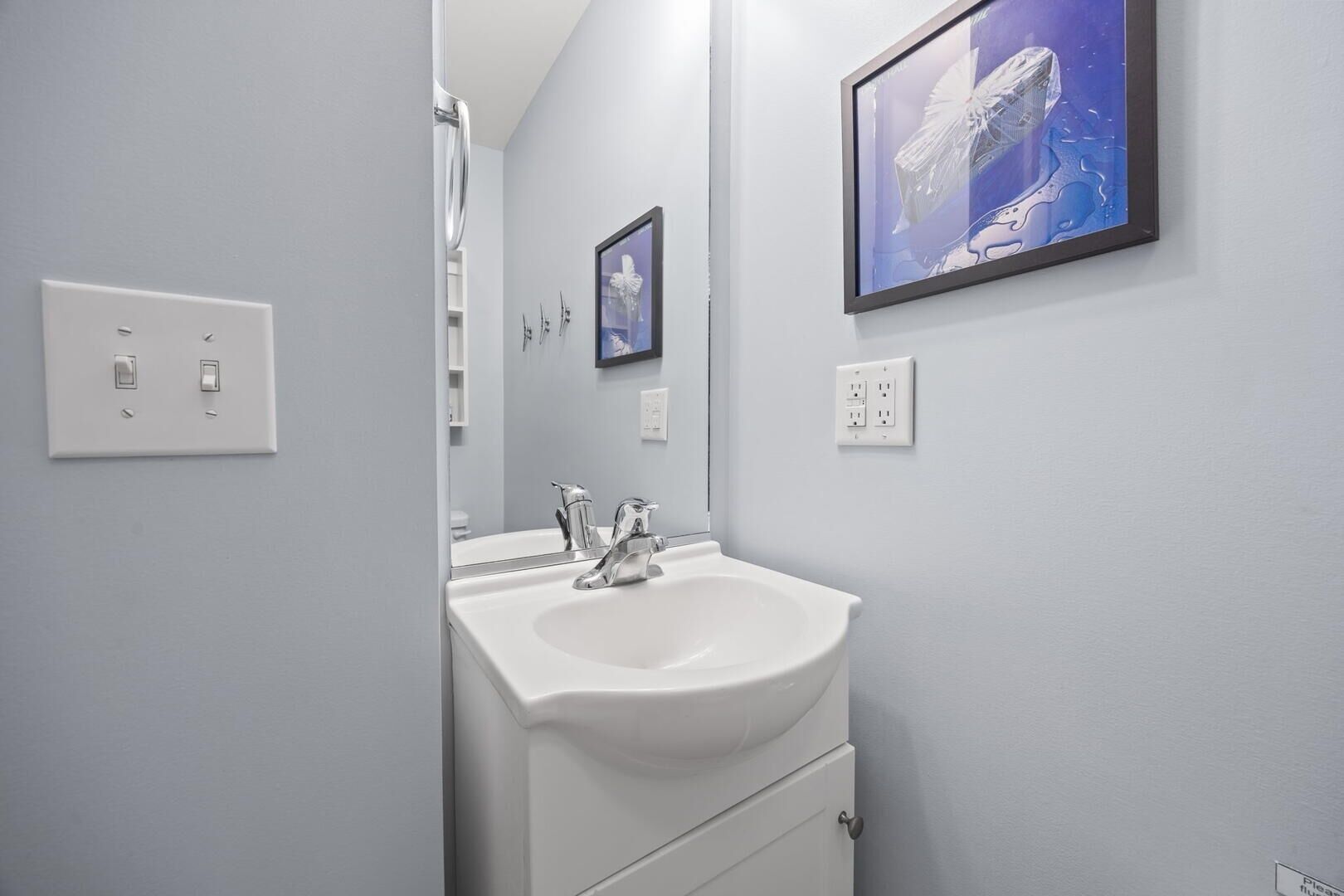 A bathroom with a white sink, a mirror, a framed picture on the wall, and a door leading to another room.