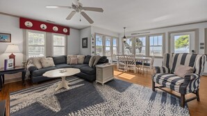 A living room with a sofa, a coffee table, a striped armchair, a dining table with chairs, a ceiling fan, and large windows. - Whispering Oaks 3 Bedroom Home by RedAwning (Harkers Island)