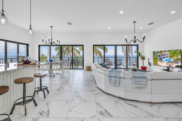 A modern living room with a white sofa, a dining area with bar stools, and large windows offering a view of the ocean.