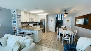 Una cocina compacta con gabinetes blancos, un microondas integrado y un refrigerador de acero inoxidable. Hay un comedor con mesa y sillas blancas, y una pequeña isla con pileta. El piso está cubierto de cerámica, y una escalera conduce a otro nivel.