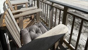 Terraza de madera con una silla acolchada y una mesa, con vista a una playa, el océano y un cielo despejado.