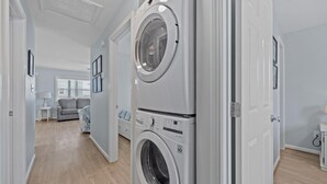 Ein Waschraum mit zwei übereinander gestapelten Frontlader-Waschmaschinen, einem grauen Sofa und einem kleinen Tisch mit einer Lampe in einem Wohnbereich.