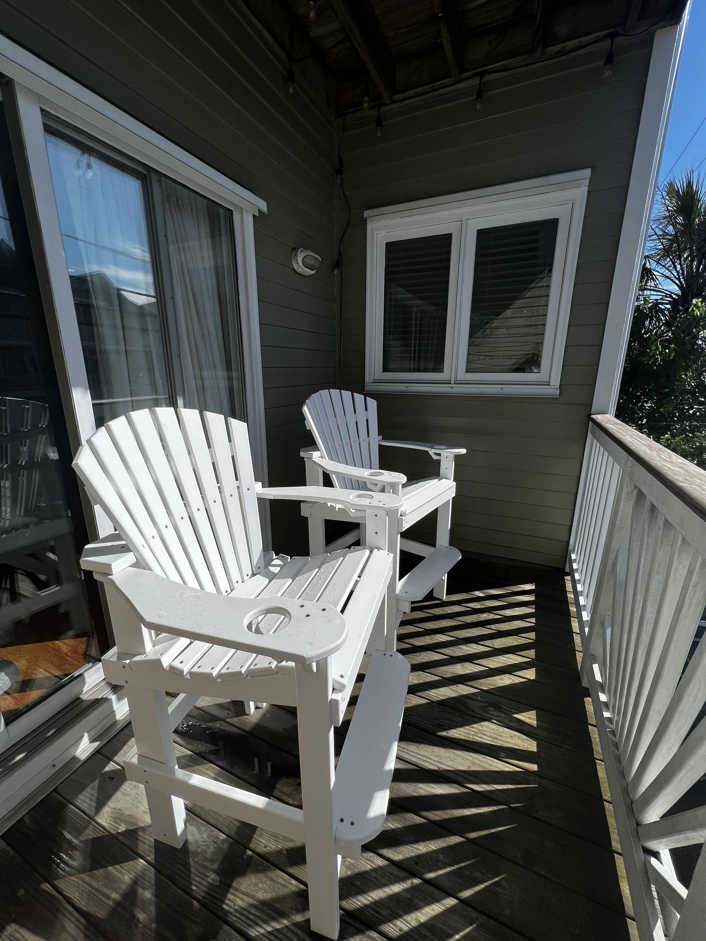 Ein überdachter Außenbereich mit zwei weißen Adirondack-Stühlen, einem kleinen Tisch, einem Fenster mit Jalousien und einer Holzterrasse.