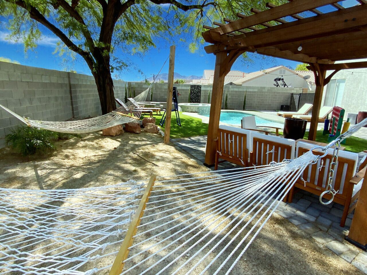 Una hamaca bajo una pérgola de madera, una piscina y un área cercada con sillones de descanso.
