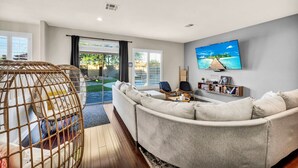 A modern living room with a grey sofa, a wicker chair, a flat-screen TV, and a bookshelf. - Buddha Estate#4 - Great neighborhood, Sparkling pool and Hot tub, EV Charger, Games (Henderson)