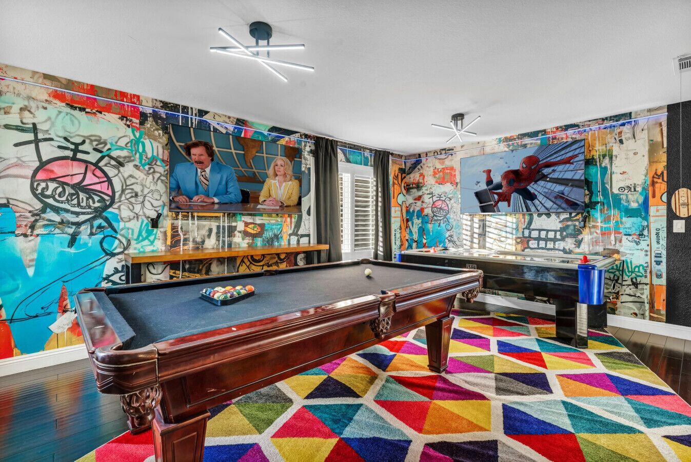 Une table de billard dans une pièce dont les murs sont couverts de graffitis et dont le sol est recouvert d’un tapis géométrique aux couleurs vives.