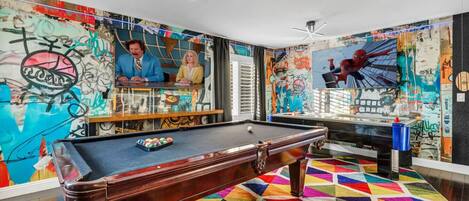 Une table de billard dans une pièce dont les murs sont couverts de graffitis et dont le sol est recouvert d’un tapis géométrique aux couleurs vives.