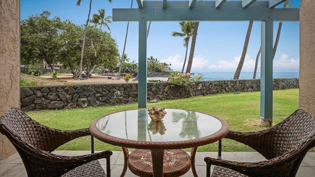 A covered patio with wicker chairs and a glass table, overlooking a tropical landscape with palm trees and a view of the ocean.