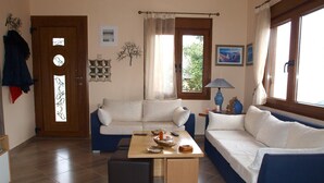 A living room with a sofa, a coffee table, and a lamp. There is a door with a glass panel and a coat rack with clothes hanging on it. - Anastasia s Cottage in Pelion (Zagora-Mouresi)