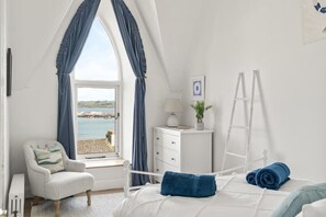 A bedroom with a bed, a chair, a dresser, a ladder, and a window with a view of the sea.