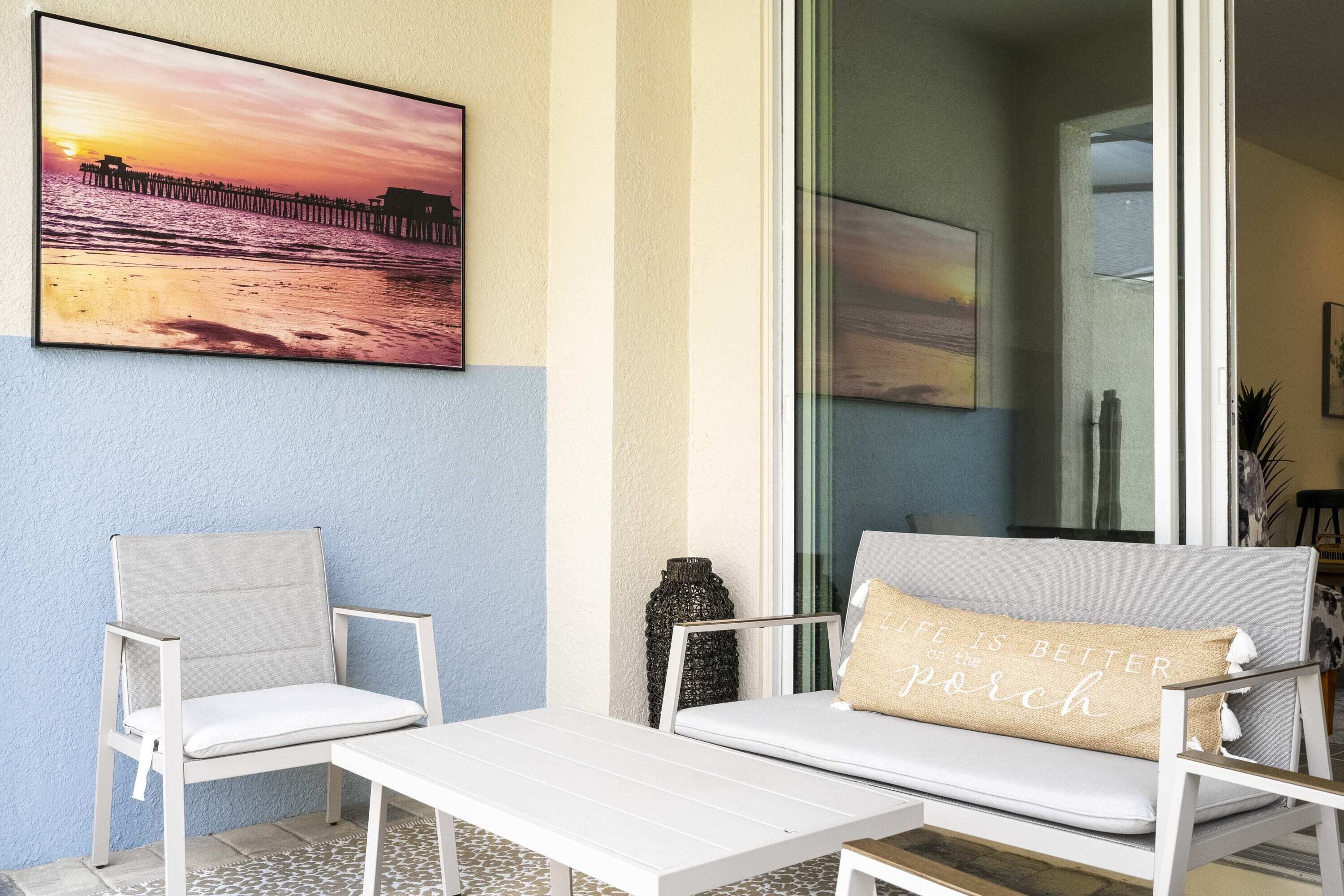 A balcony with a white sofa, a white chair, and a table. There is a painting of a sunset on the wall.