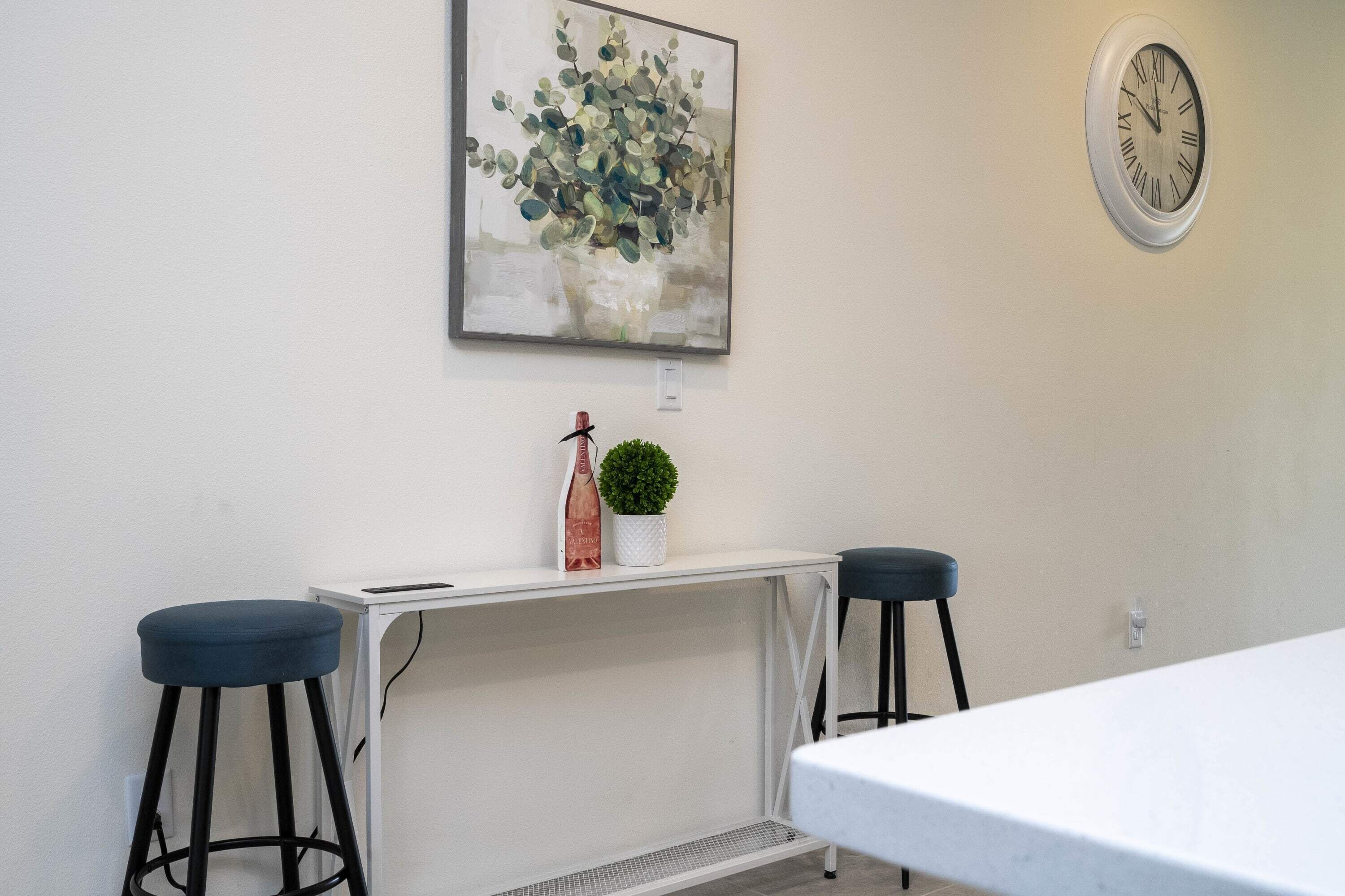 Ein Zimmer mit einem weißen Tisch, zwei schwarzen Hocker, einem weißen Beistelltisch mit Pflanze und einer Uhr an der Wand.