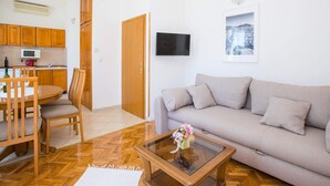 A living room with a sofa, a coffee table with a vase, a dining area with a table and chairs, wooden cabinets, a kitchen with a microwave, and a TV mounted on the wall. - Apartments Vidak - One Bedroom Apartment With Balcony and Sea View - (Konavle)