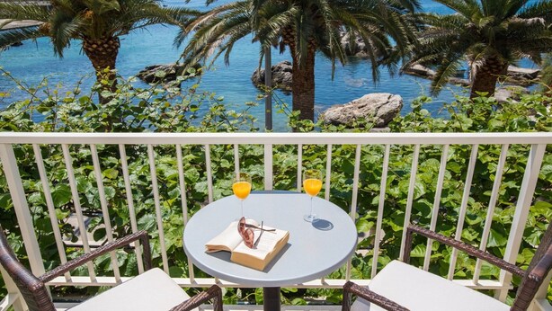 A balcony with two wicker chairs and a table, overlooking a pool with palm trees and a view of the sea. - Apartments Vidak - One Bedroom Apartment With Balcony and Sea View - (Konavle)