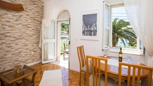 A dining area with a wooden table and chairs, a glass-top coffee table, a white rug, and a view of a palm tree and water through a window. - Apartments Vidak - One Bedroom Apartment With Balcony and Sea View - (Konavle)