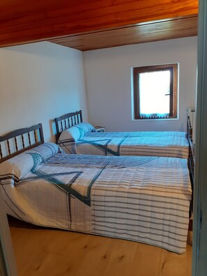 A room with two beds, wooden headboards, a window with a white curtain, and a wooden ceiling.