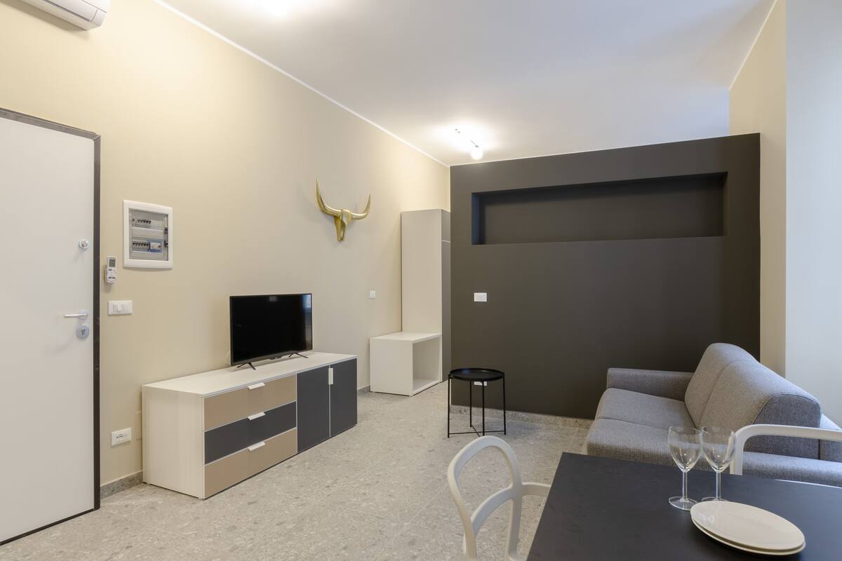 A modern living room with a sofa, a TV on a cabinet, a dining table with glasses and a plate, and a bull's head decoration on the wall.