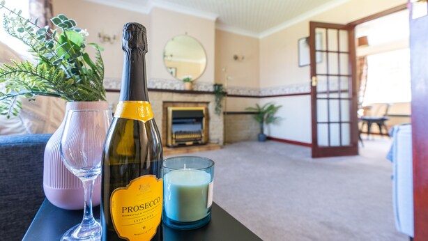A room with a fireplace, a mirror, a potted plant, and a table with a bottle, glass, and candle. - Melrose 1 (Lancaster)