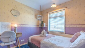 A bedroom with a bed, a desk, a chair, a bedside table, a lamp, and a window with blinds. - Melrose 1 (Lancaster)