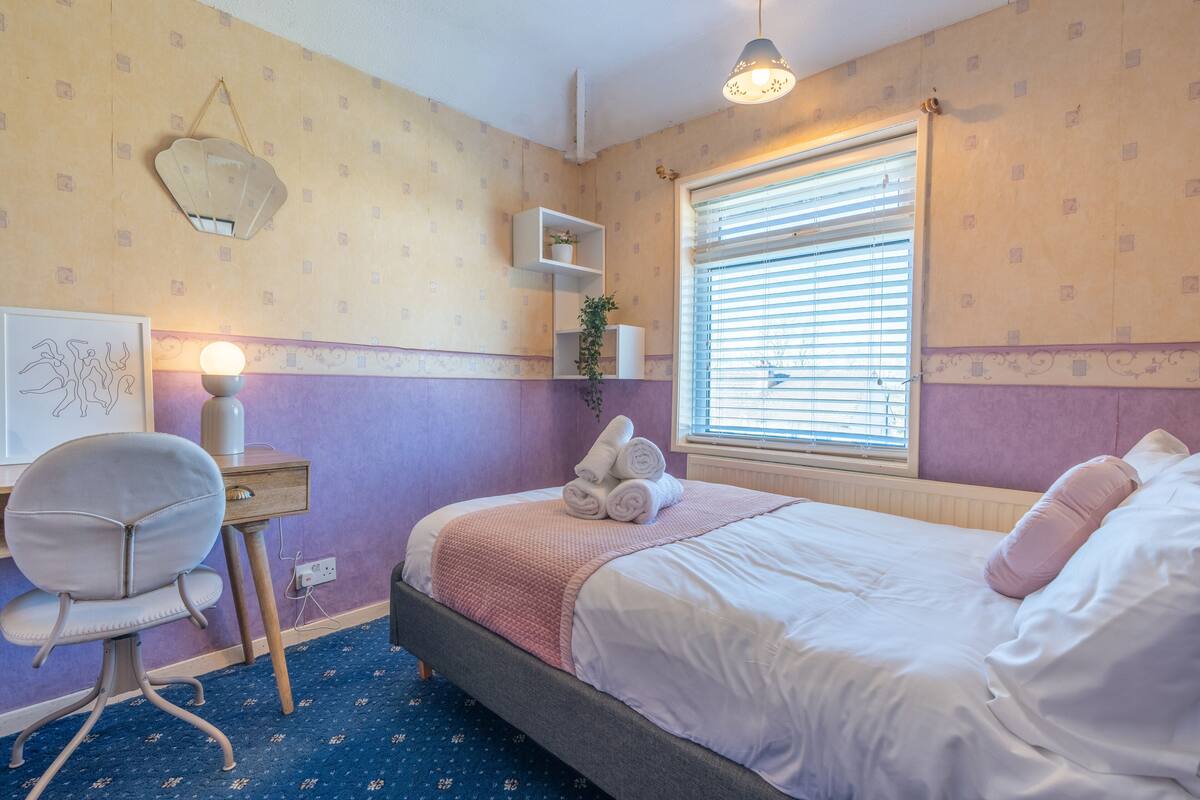 A bedroom with a bed, a desk, a chair, a bedside table, a lamp, and a window with blinds.