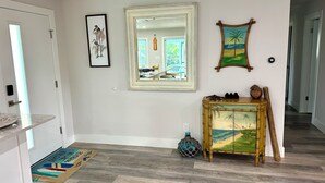 A hallway with a wooden floor, a mirror, a framed picture of birds, a decorative console table with a painting, and a doormat.