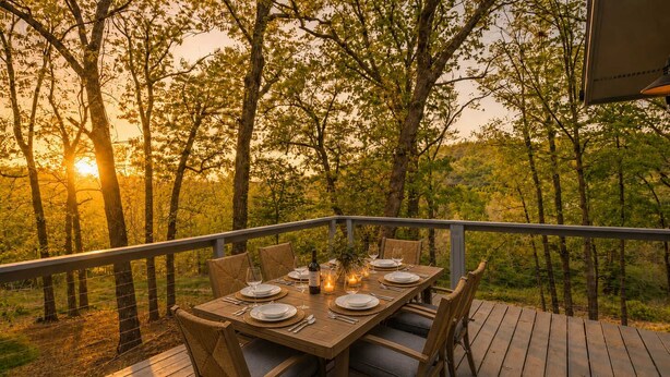 Una terraza de madera con una mesa preparada para comer, rodeada de árboles y vistas al atardecer.