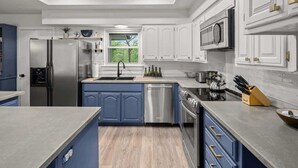 Una cocina moderna con armarios blancos, detalles azules, electrodomésticos de acero inoxidable y un amplio ventanal que proporciona luz natural.
