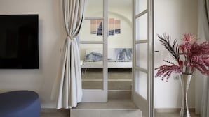 Une chambre moderne avec une grande télévision, un fauteuil bleu, une porte vitrée donnant sur un balcon et un vase rempli de fleurs roses.