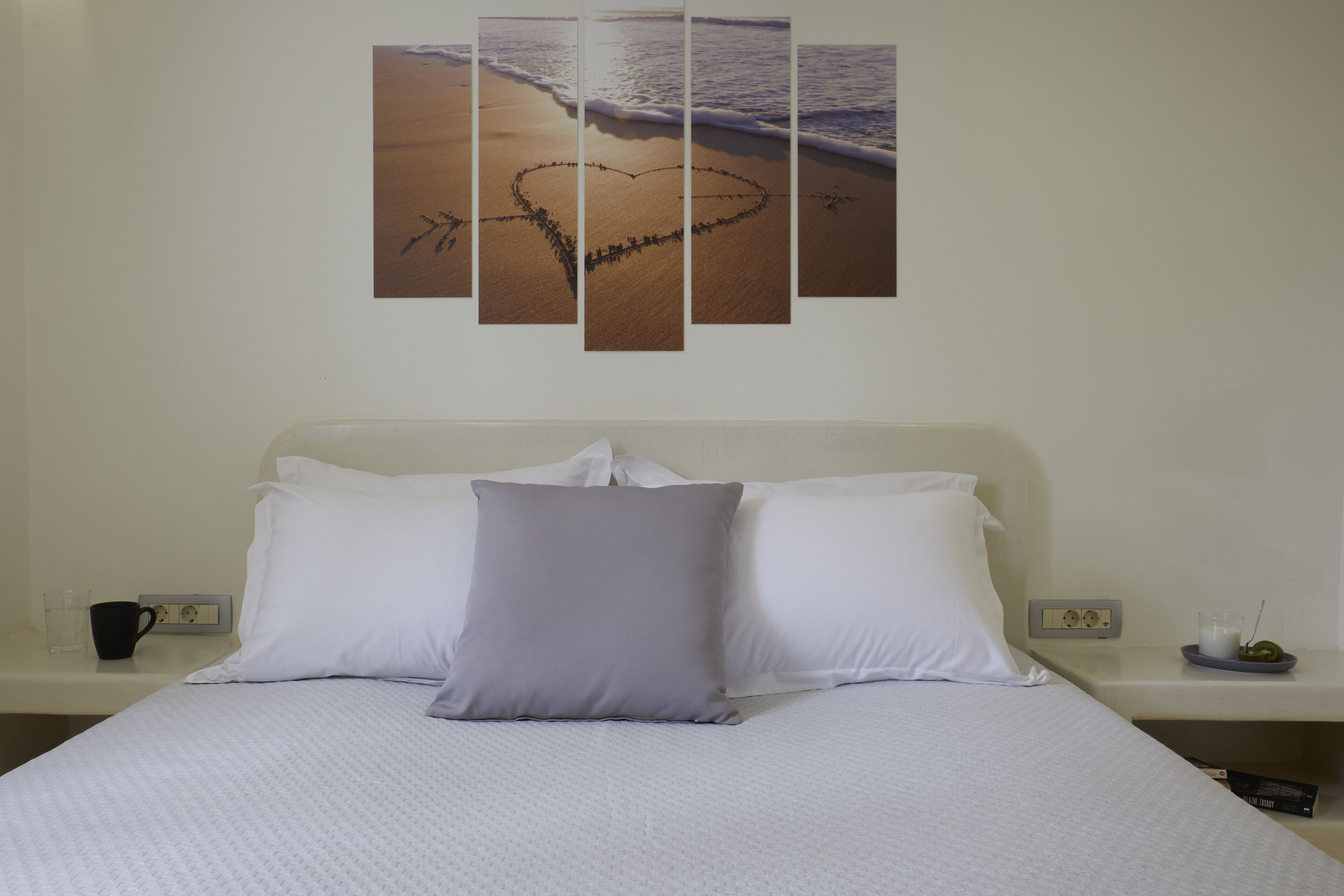 Ein ordentlich bezogenes Bett mit weißer und grauer Bettwäsche, eine herzförmige Sandzeichnung am Strand und ein an der Wand befestigtes Kunstwerk, das die Strandlandschaft zeigt.