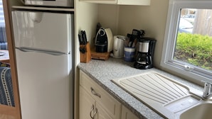 A compact kitchen with a refrigerator, microwave, coffee maker, and a window with a view of greenery. - Charmant Mobile-home Cosy (Bernières-sur-Mer)