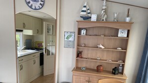 A cozy kitchen with wooden cabinets, a dining table with a pineapple-patterned tablecloth, and a small shelf with decorative items. - Charmant Mobile-home Cosy (Bernières-sur-Mer)