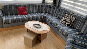 A corner seating area with a sofa and a small table, a window with blinds, and a wooden floor. - Charmant Mobile-home Cosy (Bernières-sur-Mer)