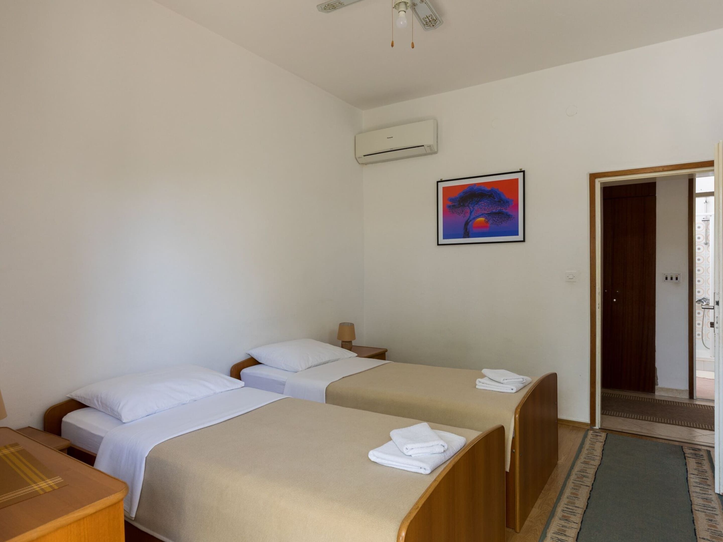 A hotel room with two single beds, a wooden desk, an air conditioning unit, and a framed painting on the wall.