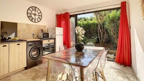 A compact kitchen with modern appliances, a dining area with a glass table, and a view of greenery through large windows. - Superbe Appartement Cosy à 300 m de la Plage (Bandol)