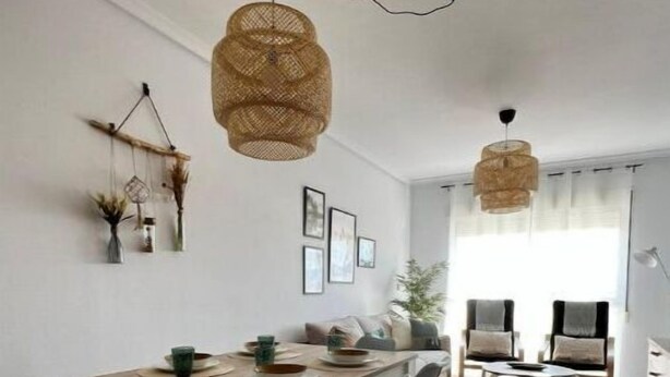 Ein moderner Essbereich mit einem Holztisch und -stühlen, einer hängenden gewebten Leuchte und Blick in ein Wohnzimmer mit Sofa und Fernseher.