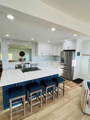 A modern kitchen with a central island, bar stools, stainless steel appliances, and a large clock on the wall. - Middle Bass Island Luxury Rental (Middle Bass)