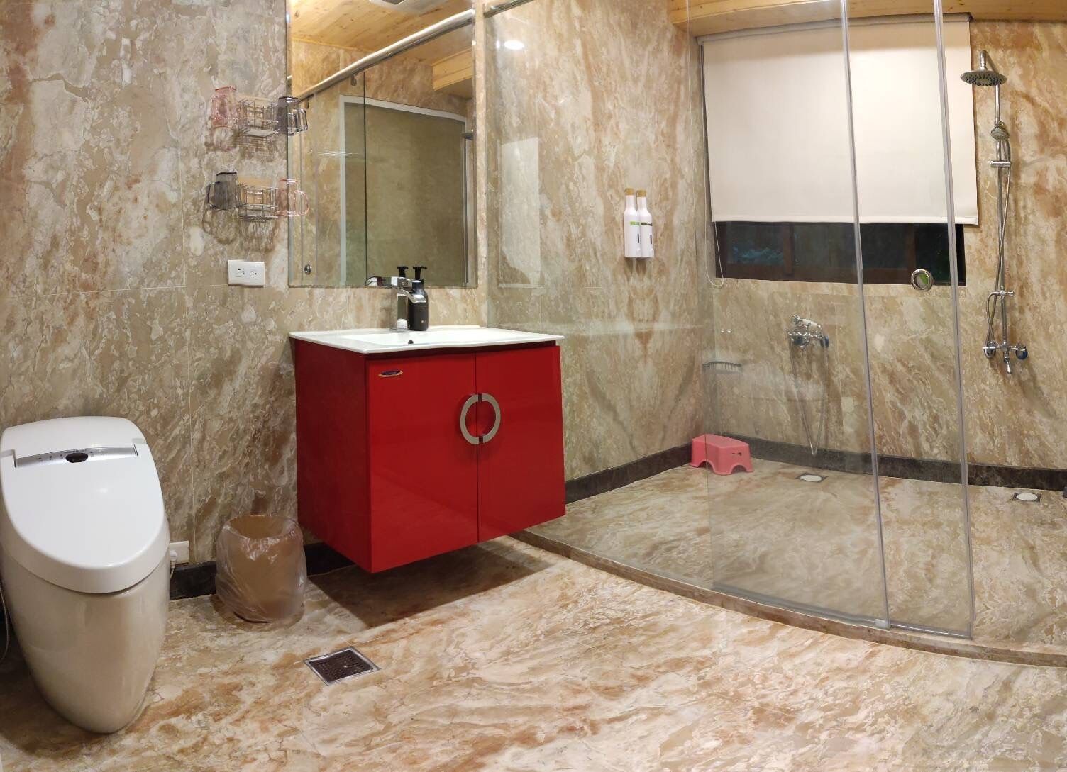 A bathroom with a glass-enclosed shower, a red vanity, a white toilet, and marble flooring.