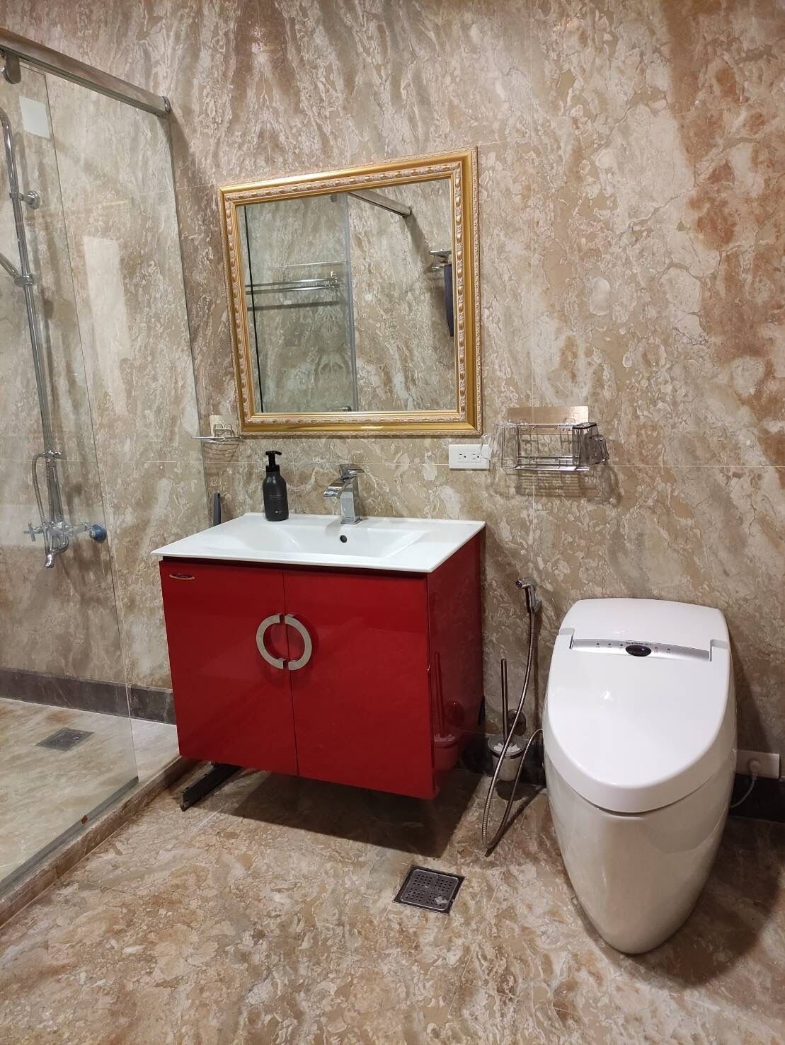 A bathroom with a marble wall, a red vanity, a white sink, a gold-framed mirror, and a white toilet.